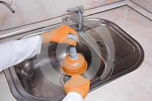 WomanÃ¢â¬â¢s hands with orange gloves cleaning sewer at kitchen faucet over metal sink. Closeup of hand with plunger